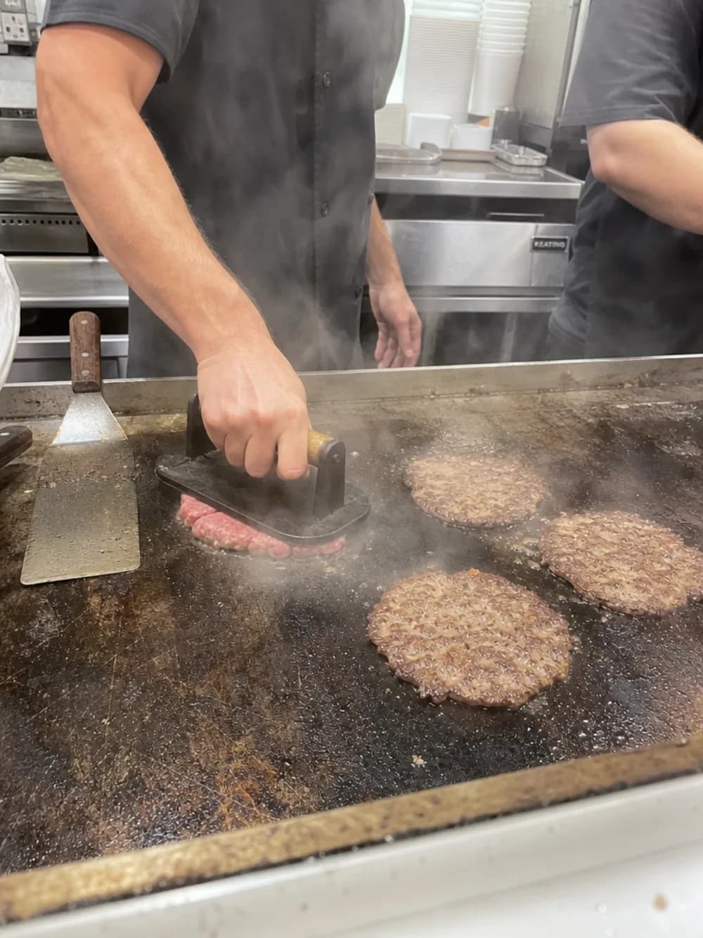 A ball of beef pressed flat on the griddle beside three searing patties